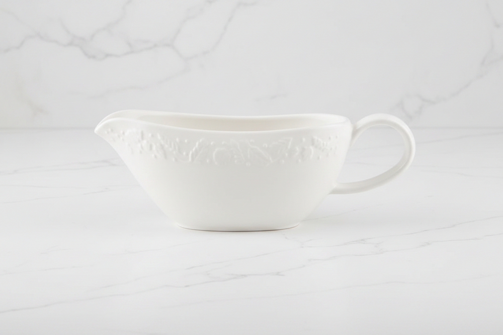 A white ceramic gravy boat with embossed autumnal floral detailing, set against a white marble background.