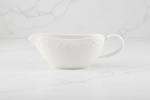 A white ceramic gravy boat with embossed autumnal floral detailing, set against a white marble background.