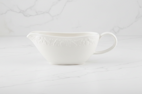 A white ceramic gravy boat with embossed autumnal floral detailing, set against a white marble background.