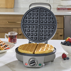 Waffle maker with waffles on a kitchen counter, surrounded by berries and tea.