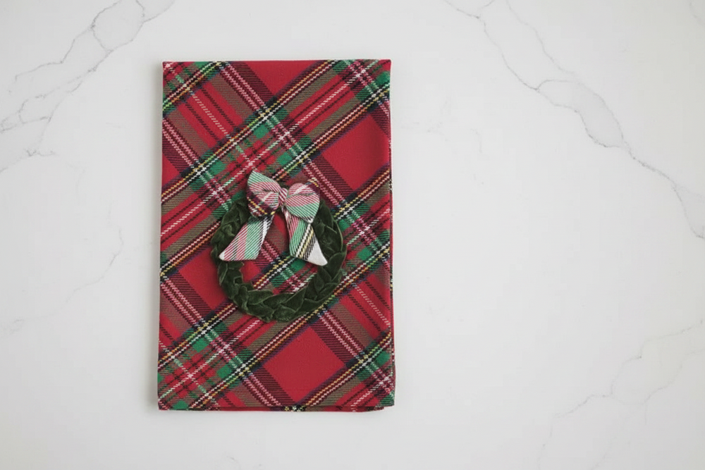 A rectangular kitchen towel with a red, green, and white plaid pattern, featuring a dark green tiered Christmas tree applique tied with a matching plaid bow at the top.