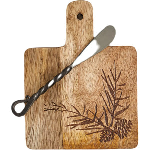 A rectangular wooden serving board with a handle and a hanging loop, featuring a dark engraved pine branch and pinecone design. A twisted metal spreader is placed diagonally across the board, which is photographed on a clean white background.