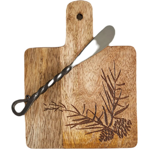 A rectangular wooden serving board with a handle and a hanging loop, featuring a dark engraved pine branch and pinecone design. A twisted metal spreader is placed diagonally across the board, which is photographed on a clean white background.