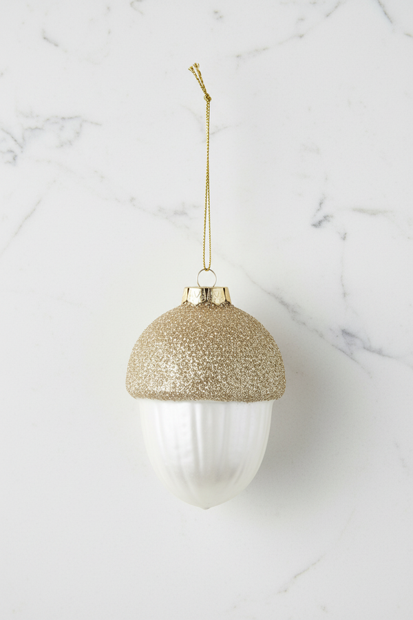 Acorn-shaped Christmas ornament with a glittery gold cap and a ribbed white body, hanging from a gold string on a white background.