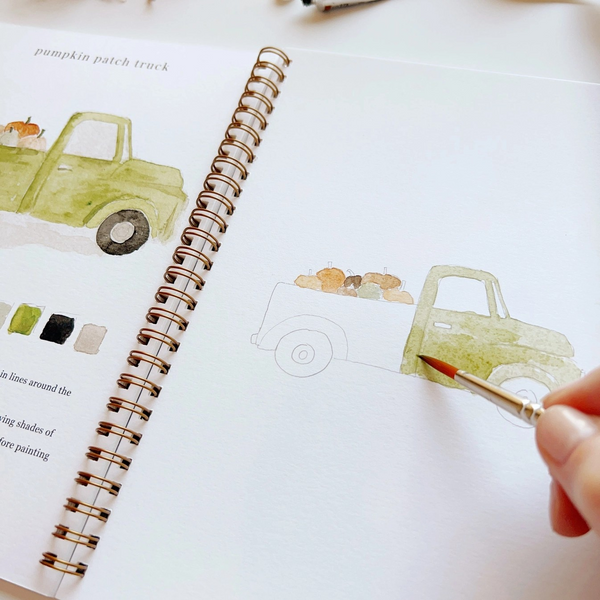 Person painting a watercolor truck illustration in a notebook with a paintbrush.