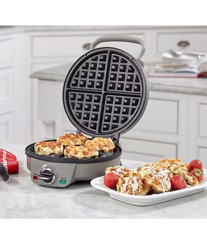 A round electric waffle maker with a gray handle and base, shown open with waffles cooking inside and a plate of finished waffles with strawberries on a marble countertop in a bright kitchen setting.