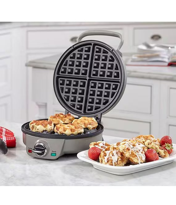 A round electric waffle maker with a gray handle and base, shown open with waffles cooking inside and a plate of finished waffles with strawberries on a marble countertop in a bright kitchen setting.