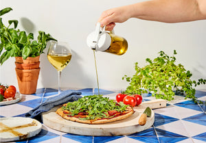 Person pouring olive oil over a pizza on a table with plants and wine.