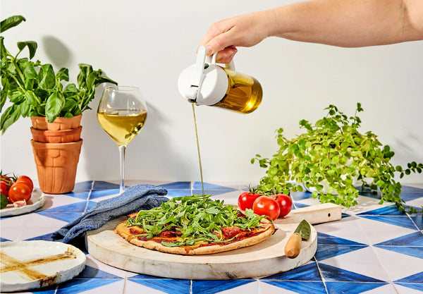 Person pouring olive oil over a pizza on a table with plants and wine.