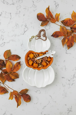 A white ceramic pumpkin-shaped dish with a hinged lid, shown open to reveal an orange-brown interior tray holding a small decorative figurine and a white spoon. The dish is photographed on a textured olive green background with autumn leaves placed around it.