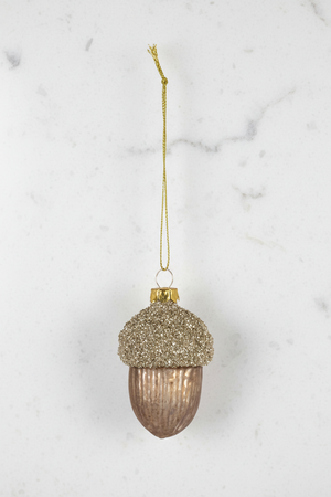 A hanging acorn-shaped ornament with a glittery gold cap and a ribbed metallic bronze-brown body, suspended by a gold string on a white background.