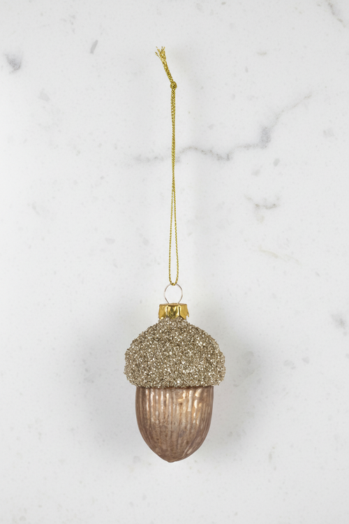 A hanging acorn-shaped ornament with a glittery gold cap and a ribbed metallic bronze-brown body, suspended by a gold string on a white background.