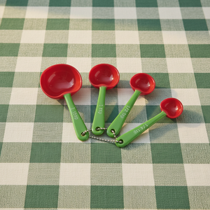 Four small cherry measuring spoons with red bowls and green handles arranged on a green and white checkered tablecloth. The handles are marked 1 tsp, 1/2 tsp, 1/3 tsp, and 1/4 tsp.