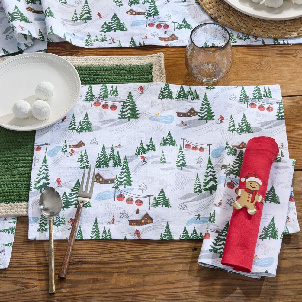 White cotton placemat with an allover ski village print of skiers, red gondolas, log cabins, ice skaters, and snowy evergreen trees.