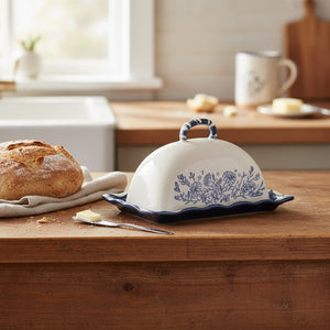 A ceramic butter dish with a domed lid, featuring a blue floral pattern on the front and a dark blue scalloped rim with a matching handle, set against a plain white background.