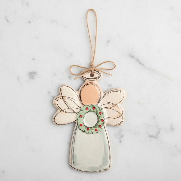 A hand-painted angel Christmas ornament with a light green dress, pale peach head, translucent wings, and a green wreath with red berries, hanging from a twine loop.
