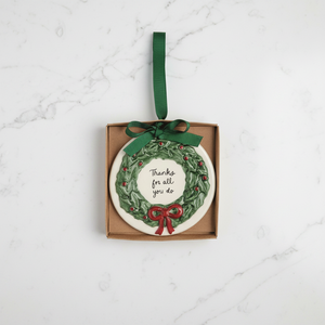 A round ceramic Christmas ornament in a square kraft gift box, featuring a green wreath design with small red berries, a green ribbon bow, and the text 'Thanks for all you do' in black script.