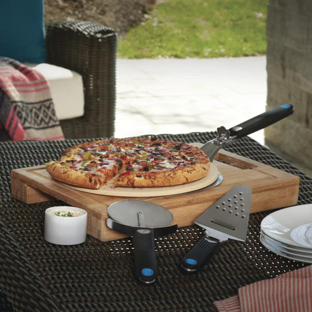 Outdoor pizza serving set on a dark woven patio table, featuring a wooden pizza board with a whole and sliced pizza, a pizza cutter, a cheese server, plates, and a small dipping dish. A wicker chair and patio background are visible.