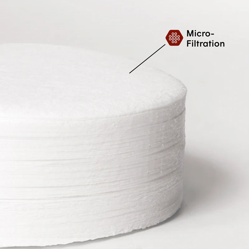 Stack of white coffee filters on a dark coaster next to a glass of black coffee on a light surface.