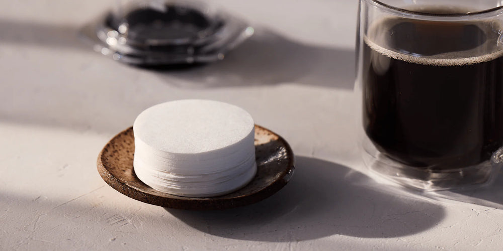 Stack of white coffee filters on a dark coaster next to a glass of black coffee on a light surface.