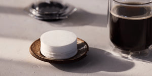 Stack of white coffee filters on a dark coaster next to a glass of black coffee on a light surface.