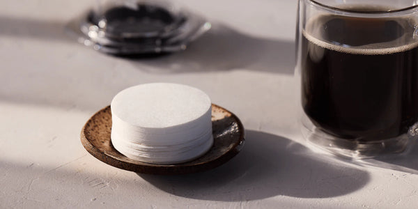 Stack of white coffee filters on a dark coaster next to a glass of black coffee on a light surface.