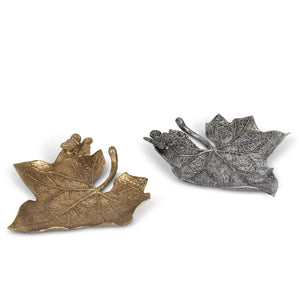 Two leaf-shaped decorative objects on a plain white background, one in metallic gold and one in metallic silver, both with textured veining and curled leaf edges.