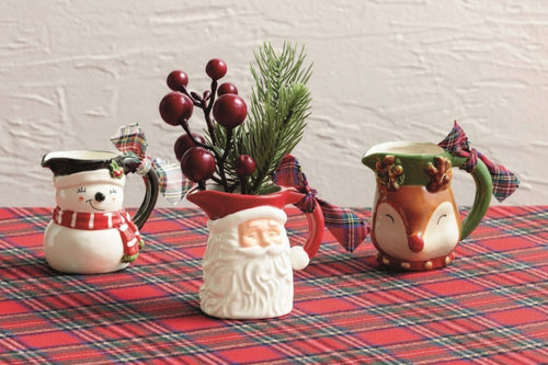 Three Christmas-themed ceramic pitchers arranged on a red plaid tablecloth against a white wall. The left pitcher is white with a snowman face and scarf; the center pitcher is white with Santa's face and holds greenery with red berries; the right pitcher is green with a reindeer face.