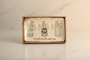 A boxed holiday gift set containing three hand-painted ceramic angels on a cream-colored background with the text 'Hark the herald angels sing' written below. The box is wrapped in twine and contains a small button with a 'Click to zoom' overlay on a solid white background.