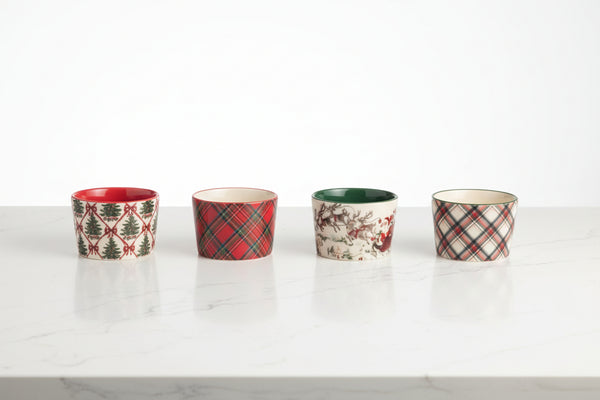 Four small ceramic cups lined up on a plain white background, each with a different holiday pattern including plaid, pine trees, and floral motifs in red, green, beige, and white.