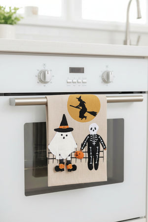 A rectangular beige fabric wall hanging or panel featuring a Halloween scene with a white ghost, a black-and-white skeleton, an orange pumpkin, and a witch silhouette against a yellow moon. A gray 'Click to zoom' button appears below the product on a white background.