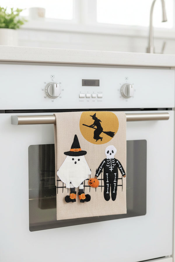 A rectangular beige fabric wall hanging or panel featuring a Halloween scene with a white ghost, a black-and-white skeleton, an orange pumpkin, and a witch silhouette against a yellow moon. A gray 'Click to zoom' button appears below the product on a white background.