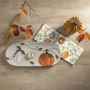 A ceramic fall-themed serving tray and matching napkin set on a textured green fabric background, styled with dried autumn leaves and decorative pumpkins. The tray features a white base with a raised orange pumpkin, a white pumpkin, and a small floral motif, accented by green leaves and vines.