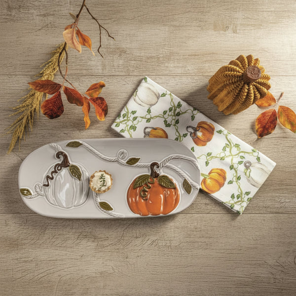 A ceramic fall-themed serving tray and matching napkin set on a textured green fabric background, styled with dried autumn leaves and decorative pumpkins. The tray features a white base with a raised orange pumpkin, a white pumpkin, and a small floral motif, accented by green leaves and vines.