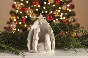 A small white nativity scene figurine with a star-topped shelter and three figures, displayed on a tabletop with green pine branches against a textured gray wall. A 'Click to zoom' overlay appears at the bottom.