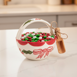 A small white ceramic Christmas basket with a red scalloped rim, green holly leaves, red berries, and a red bow, featuring a handle tied with twine to a small wooden tag.