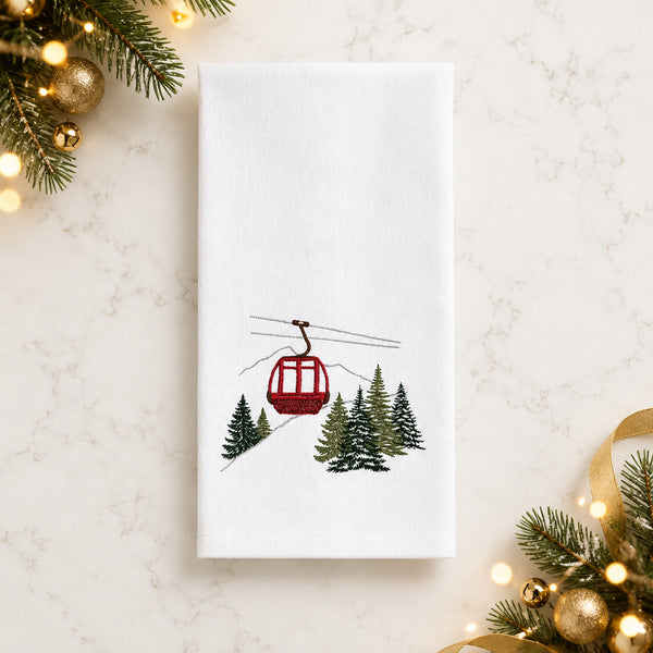White flour sack kitchen towel embroidered with a red ski gondola over snowy mountains and evergreen trees.