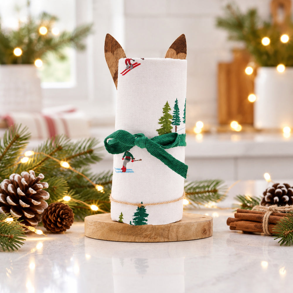 A rolled white fabric kitchen towel with a winter ski and tree print, tied with a green ribbon and displayed on a wooden stand against a white background.