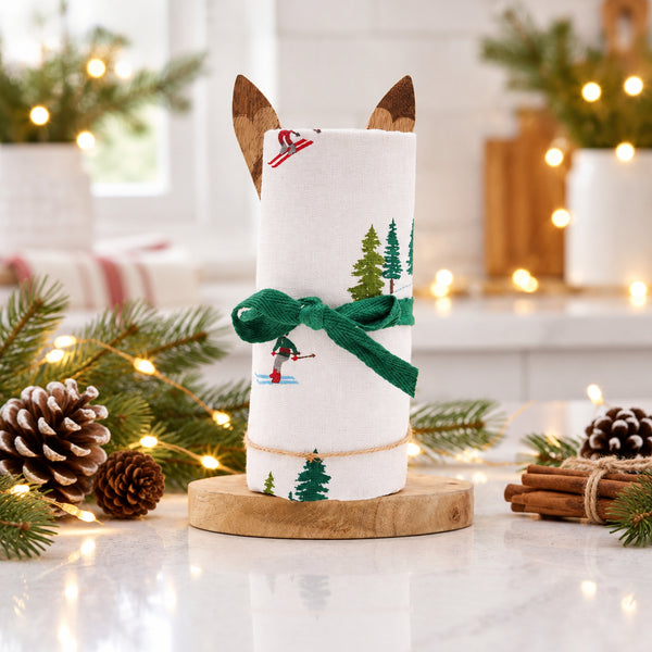 A rolled white fabric kitchen towel with a winter ski and tree print, tied with a green ribbon and displayed on a wooden stand against a white background.
