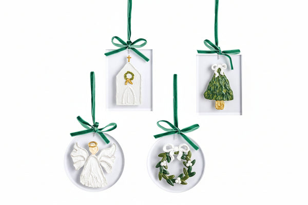 Four white Christmas ornaments with green ribbons and hanging loops, shown on a white background. The set includes a church, a Christmas tree with a light bulb, an angel, and a wreath with a bow and olive branch.