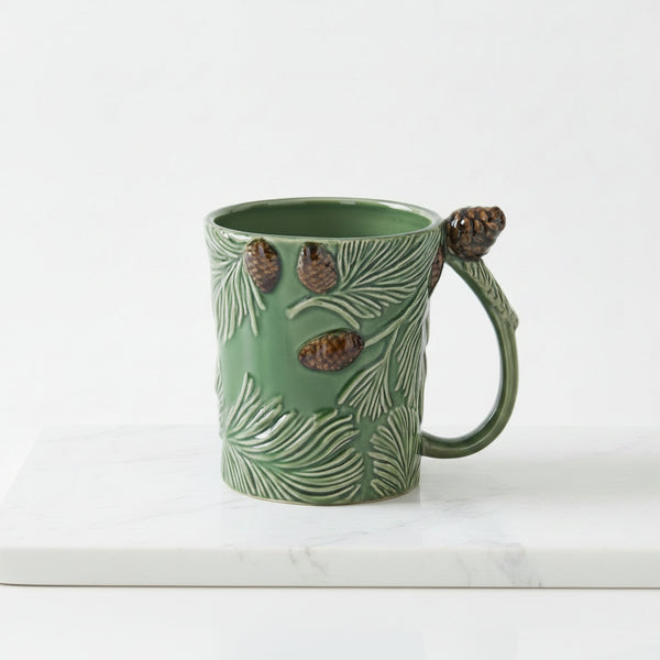 A green ceramic mug designed to look like a pine branch, featuring embossed pine needles, three raised pinecones on the body, and a pinecone attached to the handle, shown against a plain white background.