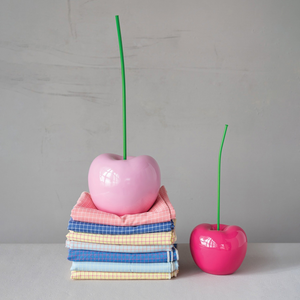 Two glossy cherry-shaped decorative objects with long green stems sit on a tabletop against a light gray wall. The larger pale pink cherry is placed on top of a stack of folded striped fabric napkins, and a smaller vivid pink cherry is beside it.