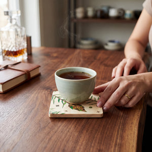 Aspen Kitchens Home+Gift ceramic coaster with pinecone design under a steaming tea cup on a rustic wooden table