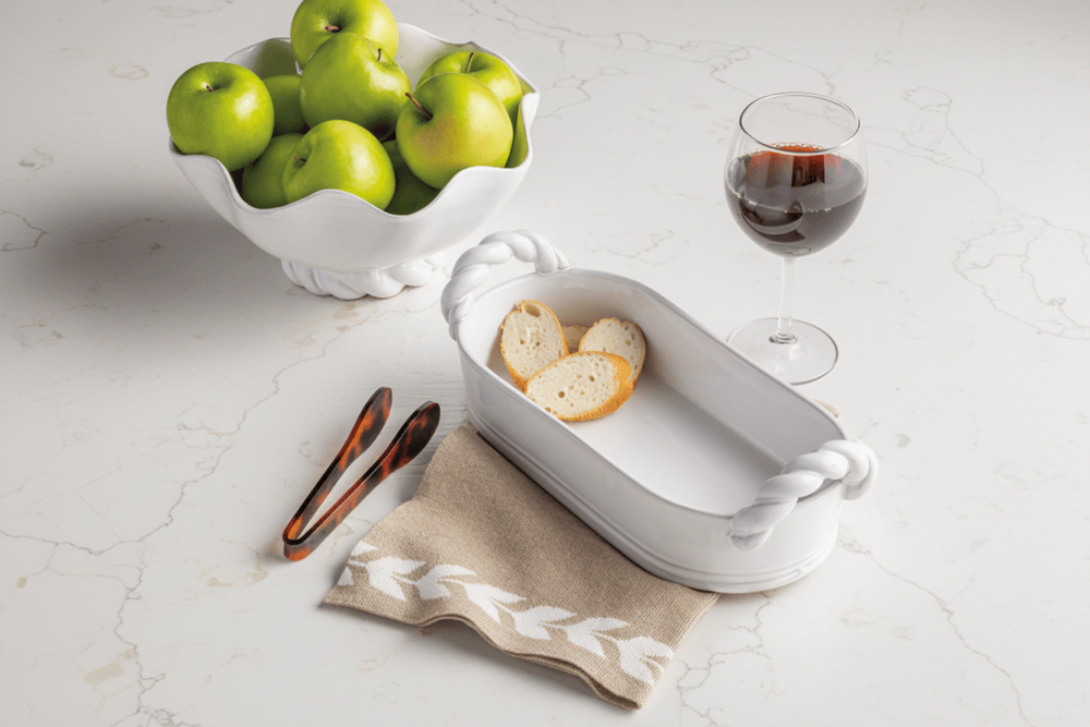 A white oval serving tray with rope-style handles sits on a beige napkin on a light textured surface, holding two slices of bread. A white bowl of green apples and a glass of red wine are placed nearby as styled props.