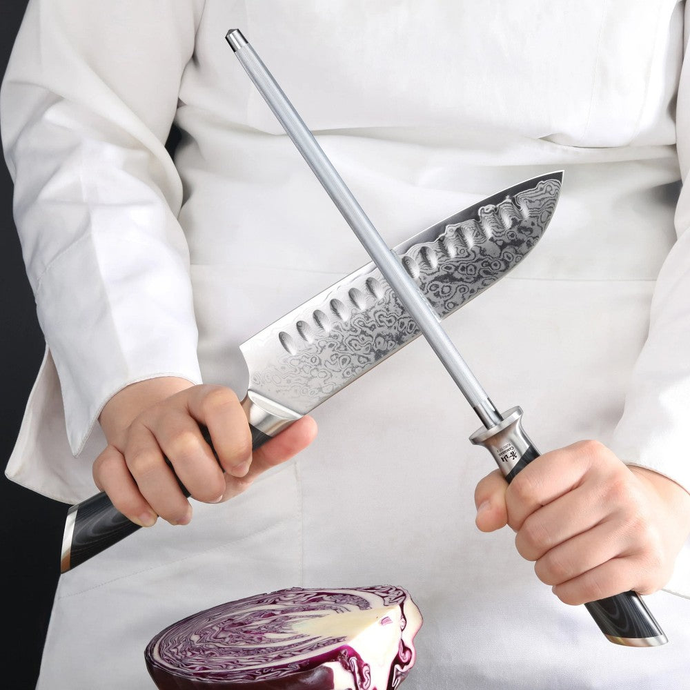 A close-up of a person in a white chef's coat sharpening a patterned Damascus-style kitchen knife with a metal honing rod, with a sliced purple cabbage visible below.