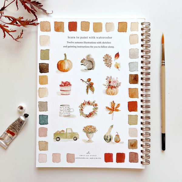 Artbook with watercolor illustrations of autumn scenes on a white surface with a paintbrush and water bottle.