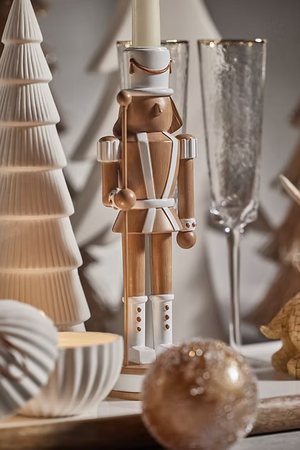 A wooden nutcracker figurine candle holder with white accents, styled on a tabletop with a textured cream Christmas tree, glass flutes, and a gold-toned ornament in a warm, softly lit setting.
