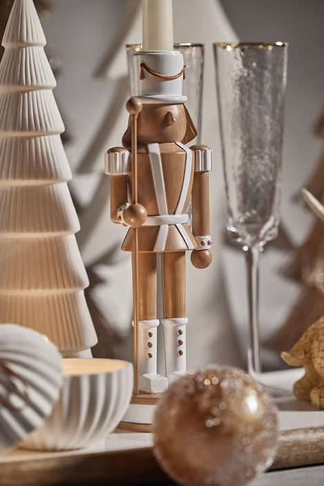 A wooden nutcracker figurine candle holder with white accents, styled on a tabletop with a textured cream Christmas tree, glass flutes, and a gold-toned ornament in a warm, softly lit setting.