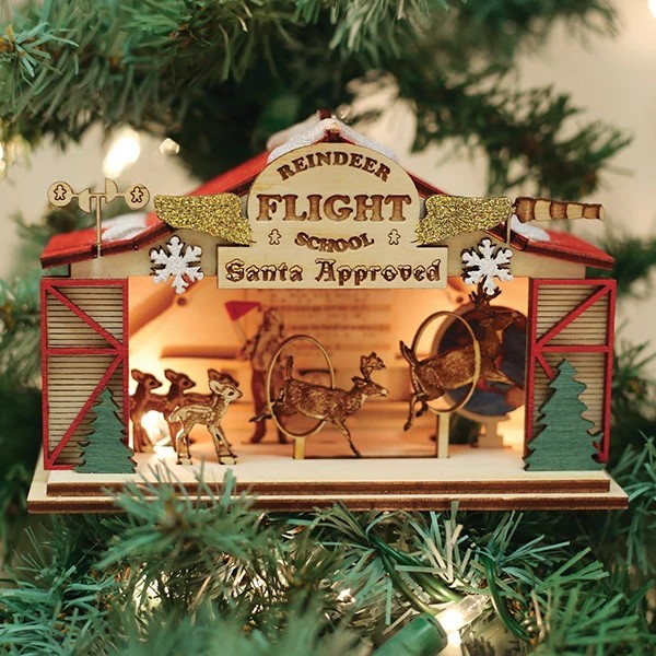 Wooden Reindeer Flight School cottage ornament with a “Santa Approved” sign, gold glittered wings, red barn doors, and reindeer leaping through hoops inside, glowing from a tree light bulb.