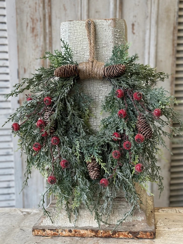 Snow-dusted balsam wreath with red frosted berries and pinecones, hanging from a jute rope loop on a weathered white door.